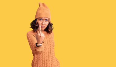 Young beautiful mixed race woman wearing wool sweater and winter hat showing middle finger, impolite and rude fuck off expression 