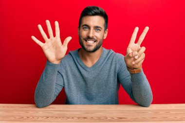 Yakışıklı İspanyol bir adam gündelik tarzda giyinmiş masada oturuyor ve yedinci parmaklarını göstererek gülümsüyor ve mutlu.. 