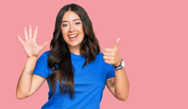 Güzel, esmer, gündelik kıyafetler giyen, altı numaralı parmaklarıyla işaret eden, kendinden emin ve mutlu bir şekilde gülümseyen genç kadın.. 