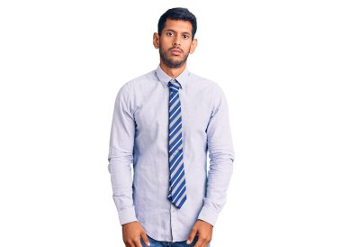 Yüzünde ciddi bir ifade olan iş kıyafetleri giyen genç bir latin. Basit ve doğal bir şekilde kameraya bakıyorsun.. 