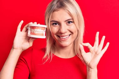 Takma dişleri olan genç, sarışın bir kadın izole edilmiş kırmızı arka planda duruyor. Parmaklarıyla işaret veriyor, gülümseyerek mükemmel bir sembol oluşturuyor.