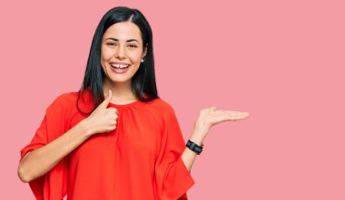 Güzel, genç bir kadın günlük giysiler giyer, avuç içini gösterir ve baş parmaklarıyla iyi bir jest yapar, mutlu ve neşeli gülümser. 