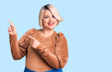 Genç sarışın, iri yarı, süveter giyen, gülümseyen ve kameraya bakan iki eli ve parmakları yandan gösteren bir kadın.. 