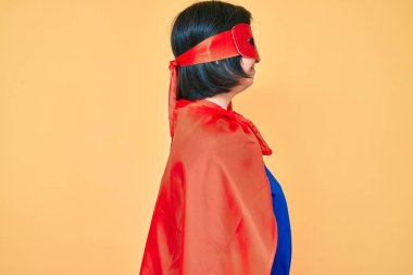 Down sendromlu esmer kadın, süper kahraman kostümü giyiyor. Yan tarafa bakıyor, rahat poz veriyor, kendine güvenen bir gülümsemesi var.. 