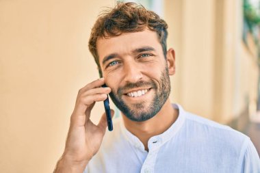 Güneşli bir günde sakallı, günlük beyaz gömlek giyen yakışıklı adam dışarıda mutlu bir şekilde gülümserken akıllı telefon kullanıyor.