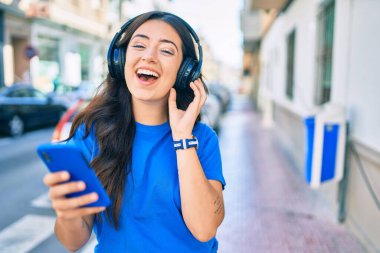 Genç İspanyol kadın şehirde akıllı telefon ve kulaklık kullanarak mutlu bir şekilde gülümsüyor..