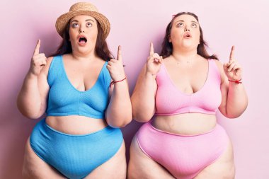 Bikini giyen genç ve cüsseli ikizler şaşkın şaşkın yukarı bakıyor ve parmaklarıyla işaret ediyor ve kollarını kaldırıyor.. 