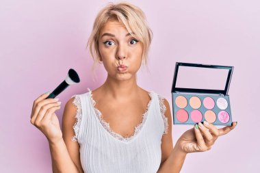 Makyaj fırçası tutan genç sarışın kız ve komik yüzüyle yanakları kızardı. Ağzı havayla şişer, havayı yakalar.. 