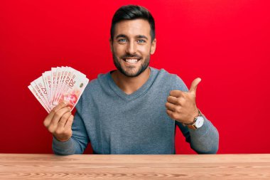 Yakışıklı İspanyol adam İsrail şekellerini tutuyor mutlu ve pozitif gülümsüyor, başparmağıyla mükemmel ve onay işareti yapıyor. 