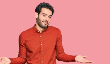 Sıradan kıyafetler giyen, elleri ve kolları havada, kafası karışmış genç İspanyol bir adam. şüphe kavramı. 