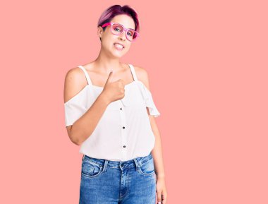 Pembe saçlı, sıradan kıyafetler giyen ve gözlüklü genç ve güzel bir kadın mutlu bir el hareketi yapıyor. Kameraya bakıp başarıyı gösteren ifade. 