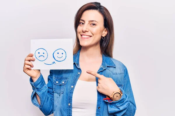 Young beautiful woman holding sad to happy emotion paper smiling happy pointing with hand and finger