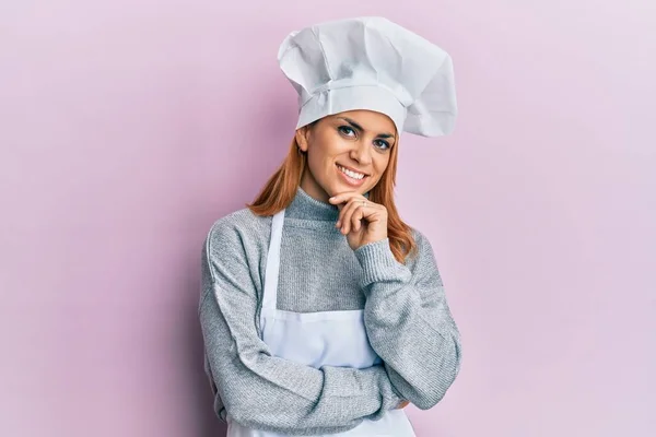 身穿职业厨师制服 戴着帽子的西班牙裔年轻女性面带微笑地看着摄像机 双手交叉 双手托着下巴 积极地思考 — 图库照片