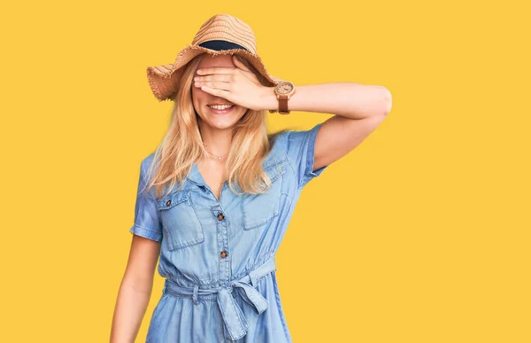 若い美しいブロンドの女性は夏の帽子をかぶって笑顔で 驚きのために目を覆う顔に手で笑っています 視覚障害の概念 — ストック写真