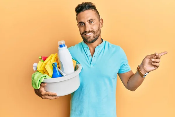 Handsome man with beard holding cleaning products smiling happy pointing with hand and finger to the side