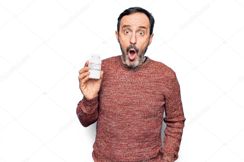 Middle age handsome man holding bottle of medicine pills over isolated white background scared and amazed with open mouth for surprise, disbelief face