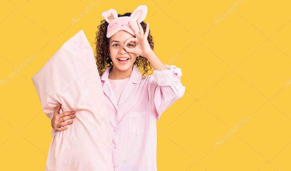 Beautiful kid girl with curly hair wearing sleep mask and pajamas holding pillow smiling happy doing ok sign with hand on eye looking through fingers 
