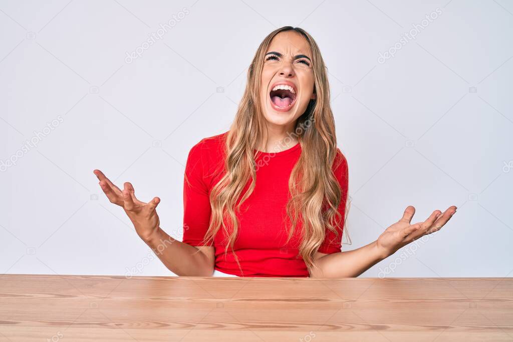 Young beautiful blonde woman wearing casual clothes sitting on the table crazy and mad shouting and yelling with aggressive expression and arms raised. frustration concept. 