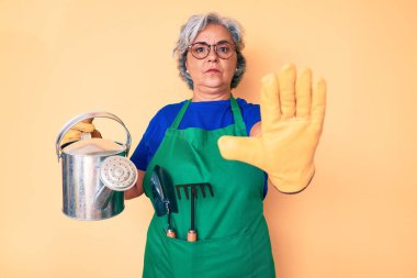 Bahçıvan önlüğü giyen, elinde su kovası tutan ve ciddi ve kendinden emin bir ifadeyle dur işareti yapan kıdemli İspanyol asıllı, kır saçlı kadın. 