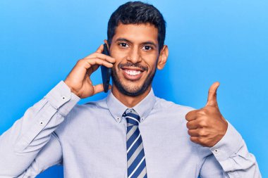 Akıllı telefondan konuşan genç Latin adam mutlu ve olumlu gülümsüyor, başparmağıyla mükemmel ve onay işareti yapıyor. 