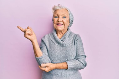 Yaşlı gri saçlı kadın yün kazak ve kış şapkası giyiyor. Gülümsüyor, mutlu bir şekilde elini ve parmağını yana uzatıyor. 