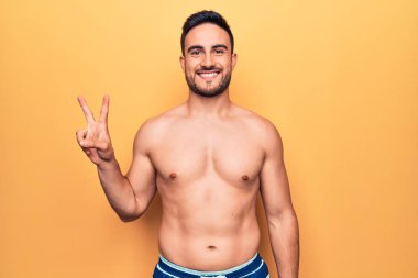 Sakallı, kolsuz tişörtlü, sarı arka planda duran ve kameraya gülümseyen, zafer işareti yapan yakışıklı genç bir adam. İki numara..