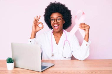 Genç Afro-Amerikan kadın doktor üniforması giyiyor elinde pembe kanser kurdelesi parmaklarıyla imza atıyor, gülümseyerek mükemmel bir sembol oluşturuyor. 