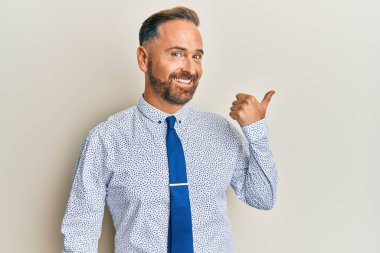Yakışıklı, orta yaşlı, iş gömleği ve kravat takan, gülen yüzlü gülümseyen ve baş parmağıyla yan tarafı gösteren bir adam.. 
