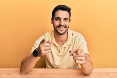 Gündelik giysiler giymiş genç İspanyol adam masaya oturmuş mutlu ve komik yüzüyle kamerayı işaret ediyor. iyi enerji ve titreşimler. 