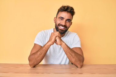 Gündelik kıyafetler giyen genç İspanyol adam masada oturmuş mutlu ve pozitif görünüyor. Kendine güvenen bir gülümsemeyle dişlerini gösteriyor. 