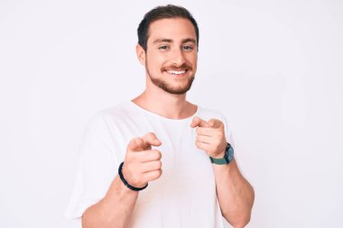 Gündelik beyaz tişört giyen genç yakışıklı adam mutlu ve komik yüzlü parmaklarını kameraya doğrultuyor. iyi enerji ve titreşimler. 