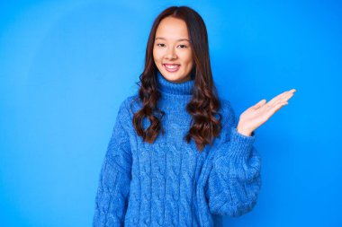 Kış süveteri giyen genç, güzel Çinli bir kız neşeli bir şekilde gülümsüyor ve avuç içi kameraya bakıyor.. 