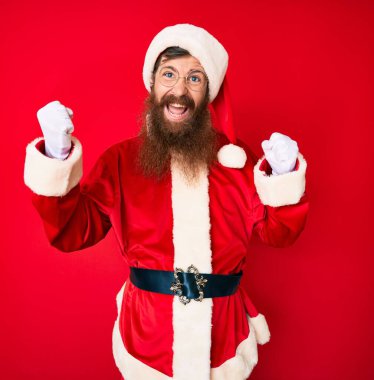 Uzun sakallı yakışıklı kızıl saçlı Noel Baba kostümü giyen gururlu, zaferi kutlayan ve kollarını kaldırarak heyecanlanan genç adam. 