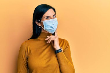 Tıbbi maske takan genç beyaz bir kadın kameraya kollarını kavuşturup çenesine dokunarak gülümsüyor. Olumlu düşünmek. 