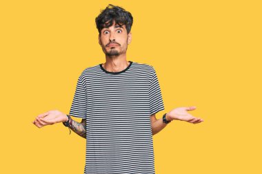 Sıradan kıyafetler giyen, elleri ve kolları havada, kafası karışmış genç İspanyol bir adam. şüphe kavramı. 