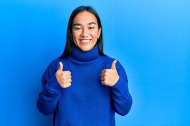 Asyalı genç bir kadın, günlük kışlık süveter giyer, başarı işareti yapar, elleriyle olumlu jest yapar, gülümser ve mutlu olur. Neşeli ifade ve kazanan jest. 
