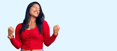 Uzun saçlı İspanyol kadın gündelik kıyafetler giyiyor. Çok mutlu ve heyecanlı. Kazanan jest yapıyor, kollarını kaldırıyor, gülümsüyor ve başarı için bağırıyor. kutlama konsepti. 