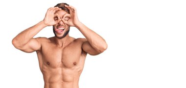 Genç beyaz adam üstsüz duruyor. Dürbün gibi jest yapıyor. Gözleri parmaklarından bakıyor. çılgın ifade. 