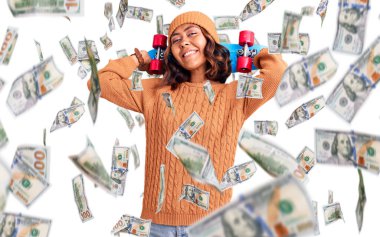 Genç, güzel, melez bir kadın elinde kaykay tutuyor. Pozitif ve mutlu görünüyor. Kendine güvenen bir gülümsemeyle ayakta duruyor ve dişlerini gösteriyor.