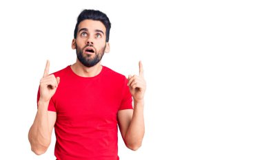 Sakallı, sıradan tişörtlü genç ve yakışıklı bir adam şaşırmış ve şaşırmış bir şekilde yukarı bakıyor ve parmaklarıyla işaret ediyor ve kollarını kaldırıyor.. 