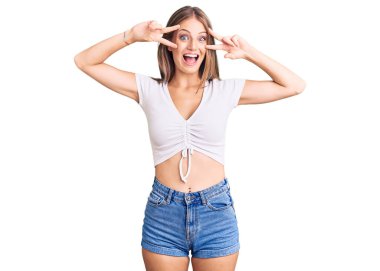 Genç, güzel, sarışın bir kadın, günlük beyaz tişört giyiyor, barış sembolü çiziyor, parmakları yüzünde, neşeyle gülümsüyor, zafer işareti yapıyor. 