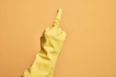 Hand of caucasian young man with cleaning glove over isolated yellow background showing provocative and rude gesture doing fuck you symbol with middle finger