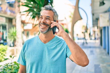 Orta yaşlı, kır saçlı, akıllı telefondan konuşan bir adam şehrin caddesinde yürüyor..