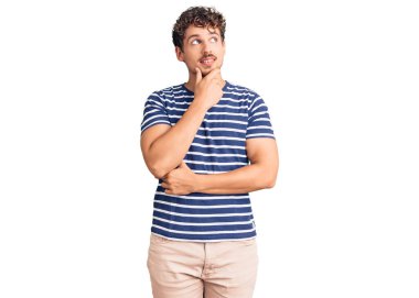 Kıvırcık saçlı, sıradan kıyafetler giyen, eli çenesinde soru soran, düşünceli bir genç adam. Düşünceli bir yüzle gülümsüyor. şüphe kavramı. 