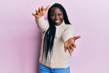 Saçları örgülü genç siyahi bir kadın sıradan bir kazak giyiyor ve kameraya sarılıp gülümsüyor. Mutluluğu kucaklayan neşeli bir ifade.. 