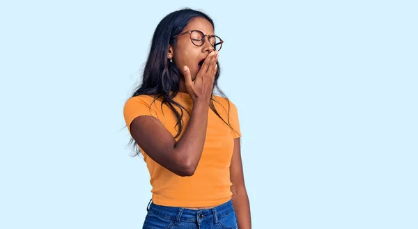 Young indian girl wearing casual clothes and glasses bored yawning tired covering mouth with hand. restless and sleepiness.