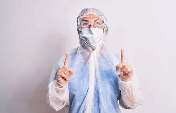 中年看護師の女性身に着けています保護コロナウイルス装置上の白い背景驚きと驚きを見上げと指差し指と上げ腕 — ストック写真