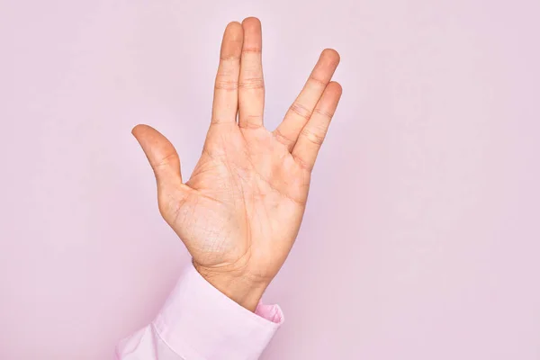 Hand of caucasian young man showing fingers over isolated pink background greeting doing Vulcan salute, showing hand palm and fingers, freak culture