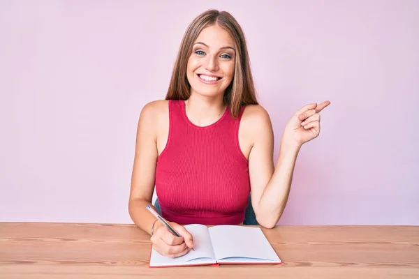 Joven Chica Caucásica Escribiendo Cuaderno Sentado Mesa Sonriendo Feliz Señalando — Foto de Stock