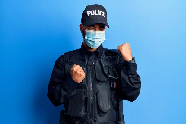 警察の制服を着た若いヒスパニック系の男と驚くべきを祝う医療マスクと腕を上げて成功のために驚いて目を閉じて — ストック写真